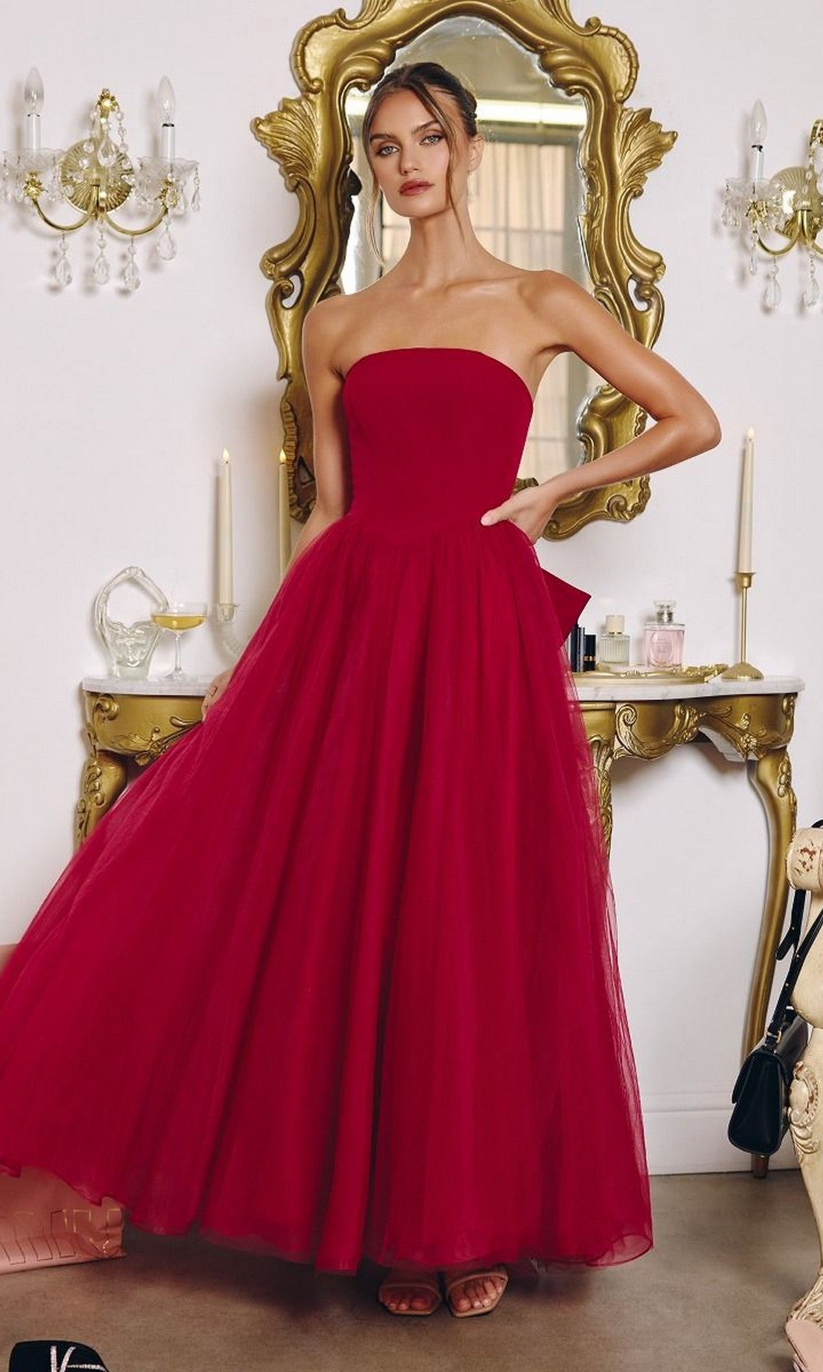 A woman stands indoors in front of a gold mirror and vanity, wearing the Classic Strapless Prom Ball Gown: Amoris T712 with a full tulle skirt.